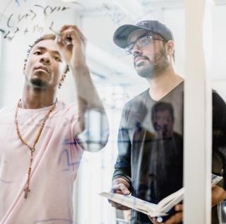 One male student holding a book looks at what another male student is writing on a glass wall